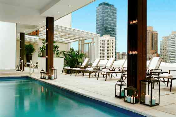 En moderne udendørs pool med solsenge og tændte lygter i et bymiljø. (AI-genereret tekst)