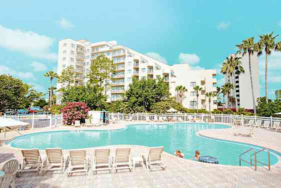 En udendørs swimmingpool foran en hvid bygning omringet af træer og liggestole, med mennesker i vandet og en klar blå himmel over. (AI-genereret tekst)