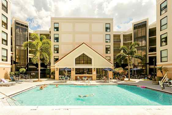 Billede af et hotel med en swimmingpool i forgrunden. Personer svømmer og slapper af ved poolområdet. Hotellets facade ses i baggrunden med palmer i nærheden. (AI-genereret tekst)