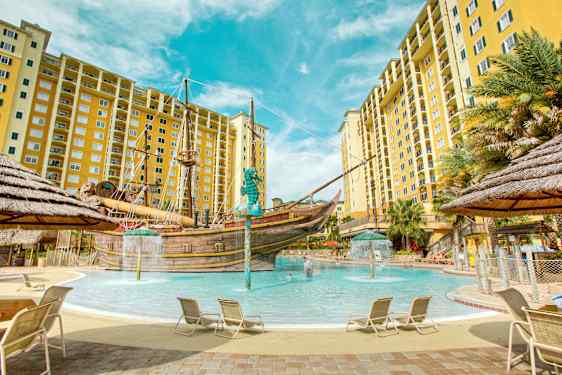 En pool omgivet af liggestole og gule lejlighedsbygninger med et piratskib i midten. (AI-genereret tekst)