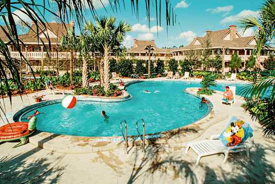 En udendørs pool omgivet af palmer og liggestole, med personer der svømmer og leger i vandet. Bygninger ses i baggrunden. (AI-genereret tekst)