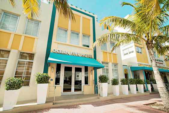 En hotelfacade med turkis baldakin og gule detaljer omgivet af palmer. (AI-genereret tekst)