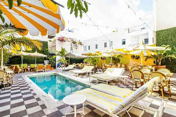 En lys og farverig udendørs pool med omkringliggende solsenge, parasoller og siddepladser omgivet af grønne planter og hvide bygninger. (AI-genereret tekst)