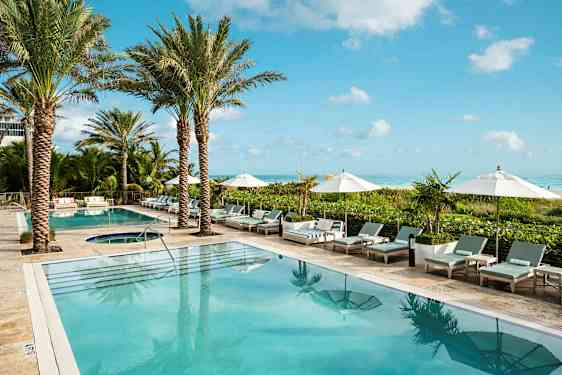 Billede af et hotel swimmingpoolområde med palmer, solsenge og parasoller ved kanten. Havet ses i baggrunden. (AI-genereret tekst)