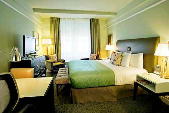 Et hotelværelse med en stor seng, et skrivebord og et tv. Værelset er indrettet i grønne toner og har store vinduer. (AI-genereret tekst)