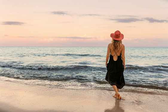 En person med en rød hat og sort kjole går på en sandstrand ved havet under en pastelfarvet himmel. (AI-genereret tekst)