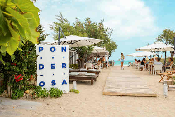 En restaurant ved stranden med borde og parasoller, en træsti fører mod vandet. (AI-genereret tekst)