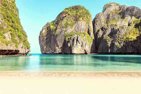 En sandstrand der vender mod roligt blåt vand, omgivet af høje klipper dækket af vegetation. (AI-genereret tekst)