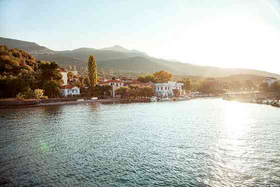 En lille landsby ligger ved en vandkant med huse og træer omgivet af bjerge. Solen skinner over området. (AI-genereret tekst)