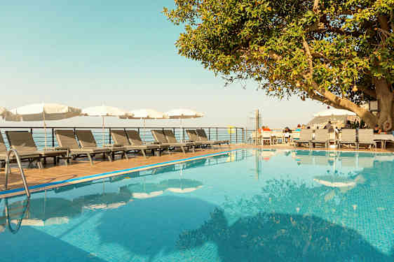 En udendørs pool med liggestole og parasoller ved siden af samt et træ og en træterrasse med siddepladser. (AI-genereret tekst)