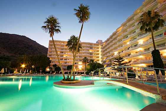 Et billede af et hotelområde ved skumring, med en oplyst pool, en bro og høje bygninger i baggrunden. (AI-genereret tekst)