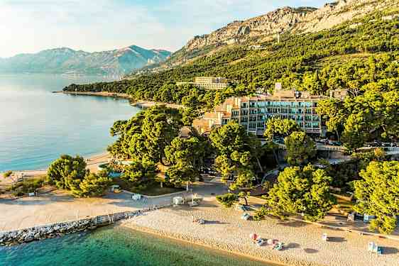 Luftfoto af en strand med parasoller, bygninger omgivet af træer og bjerge i baggrunden. (AI-genereret tekst)