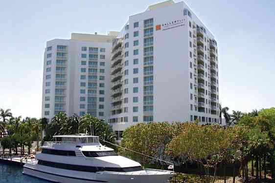 Billede af en stor hvid bygning med flere etager og altaner. Foran bygningen ses en hvid motorbåd fortøjet ved vandet samt træer og grønne områder. (AI-genereret tekst)