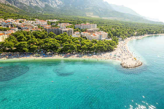 Luftfoto af en kyst med klart blåt vand, strand med liggestole og en tæt bebyggelse omkranset af skov og bjerge. (AI-genereret tekst)