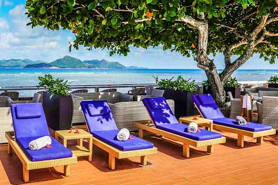 Solsenge med blå hynder og sammenrullede håndklæder placeret ved en havudsigt med bjerge i baggrunden. (AI-genereret tekst)