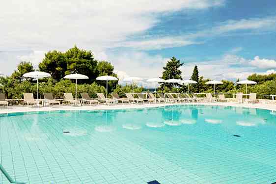 En udendørs swimmingpool omgivet af solsenge og hvide parasoller, med grøn vegetation i baggrunden og en blå himmel ovenover. (AI-genereret tekst)