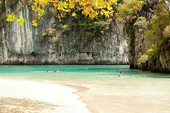 Billedet viser en strand med klart turkis vand omgivet af høje klippeformationer dækket af vegetation. Flere personer svømmer i vandet under en klar himmel. (AI-genereret tekst)