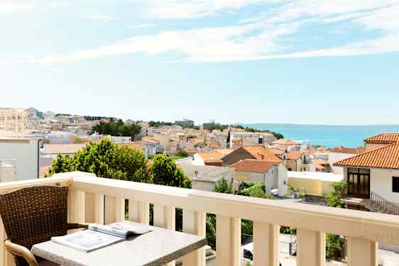 Altan med bord og stol, udsigt over en by med røde tage og havet i baggrunden. (AI-genereret tekst)