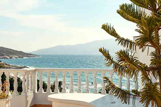 Udsigt over havet fra en terrasse med hvidt gelænder og en del af et bord i forgrunden sammen med en nåletræslignende plante. (AI-genereret tekst)