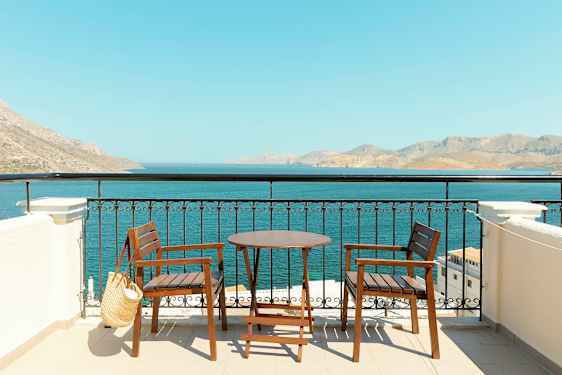 To træstole og et bord på en terrasse med udsigt til havet og bjerge i det fjerne. (AI-genereret tekst)