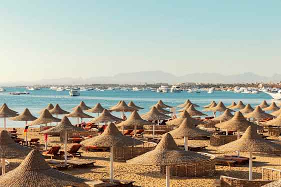 En strand med liggestole og parasoller af naturmaterialer, med havet og både i baggrunden. (AI-genereret tekst)