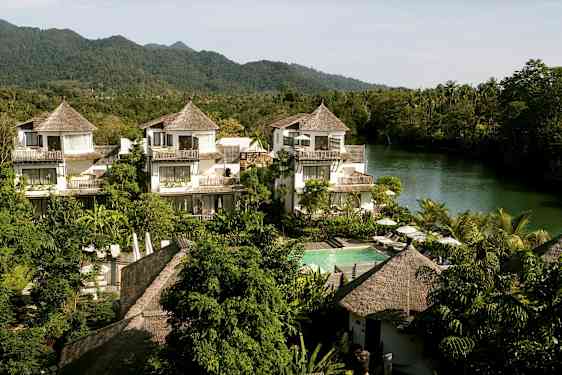 Et hotelområde med huse i tropisk stil, omgivet af grønt og en flod i baggrunden. (AI-genereret tekst)