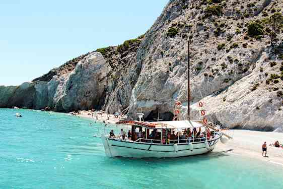 En båd ligger fortøjet ved en strand med klipper og klart turkisblåt vand. (AI-genereret tekst)