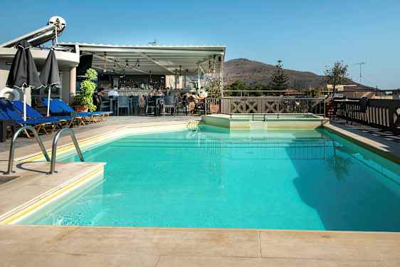 En udendørs swimmingpool med liggestole og parasoller ved siden af samt en bar i baggrunden. Et bjerg ses i det fjerne. (AI-genereret tekst)