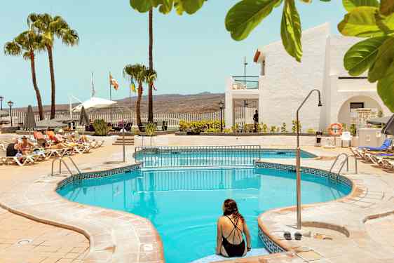 Udendørs swimmingpool med liggestole og parasoller i nærheden, samt palmer og bygninger i baggrunden. (AI-genereret tekst)