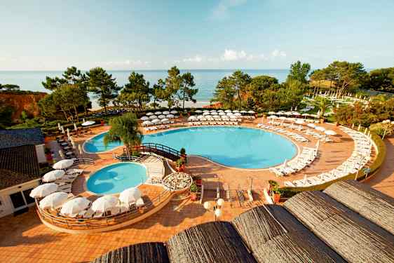 Udsigt over en swimmingpool med liggestole og parasoller, omgivet af grønne områder og med havet i baggrunden. (AI-genereret tekst)