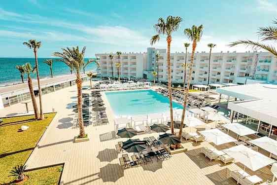 En udsigt over et feriested med en stor pool, omgivet af liggestole og parasoller, med palmer og en bygning i baggrunden tæt på havet. (AI-genereret tekst)