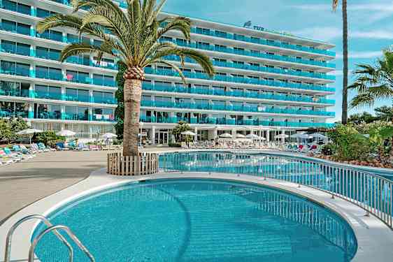 Billede af et hotel med en pool, omgivet af liggestole og parasoller, med palmer og grønne planter. (AI-genereret tekst)