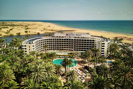 Palm Beach hotel med pool i forgrunden, omgivet af palmer, med sandklitter og hav i baggrunden. (AI-genereret tekst)