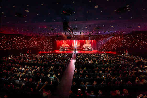 En koncertsal med en oplyst scene, hvor forskellige musikere optræder foran et publikum. (AI-genereret tekst)