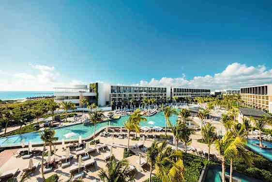 Et moderne hotelkompleks med flere pools, frodig vegetation og palmer, beliggende ved kysten med udsigt over havet under en klar blå himmel. (AI-genereret tekst)