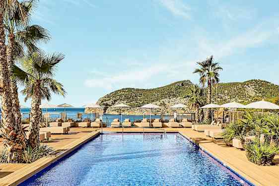 En pool med liggestole og parasoller omgivet af palmer med udsigt til havet og bjergene i baggrunden. (AI-genereret tekst)