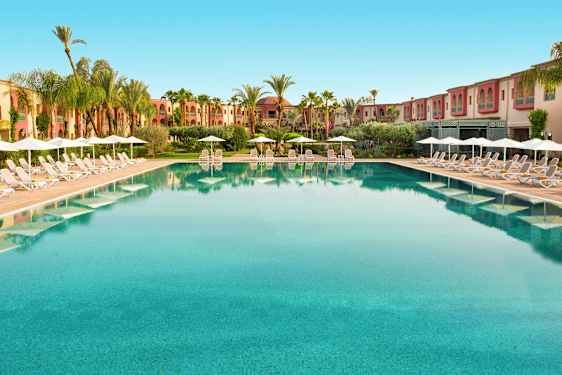 Stor pool med liggestole og parasoller langs kanten, omgivet af palmer og bygninger. (AI-genereret tekst)