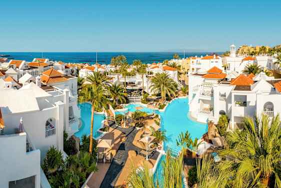 Poolområde med hvide bygninger og røde tage, omgivet af palmer med udsigt over havet. (AI-genereret tekst)