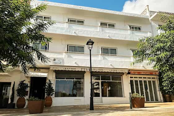 Hvid bygning med flere vinduer og altaner, omgivet af grønne områder. (AI-genereret tekst)