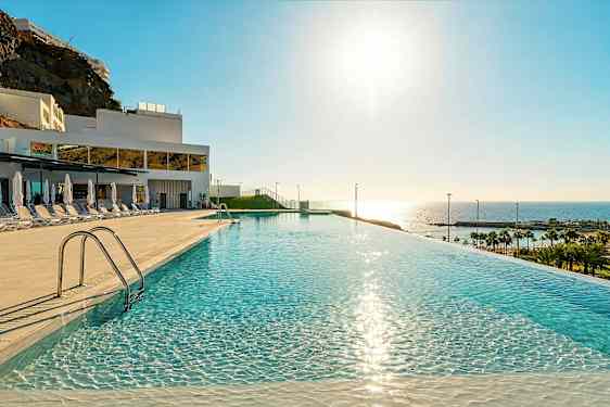 En infinitypool med spejlklart vand, solsenge og parasoller på siden, i et hotelområde med udsigt til havet og stranden. (AI-genereret tekst)