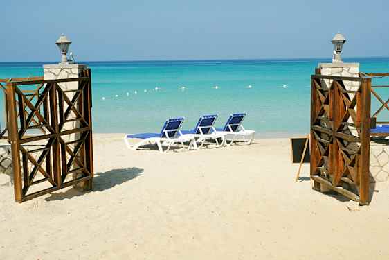 En sandstrand med liggestole og en åben port med udsigt over det turkise hav. (AI-genereret tekst)