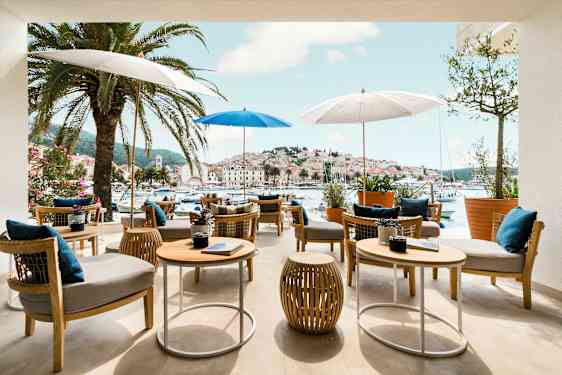Udendørs spiseområde med borde og stole under parasoller, omgivet af palmer med udsigt over en malerisk havn og by. (AI-genereret tekst)