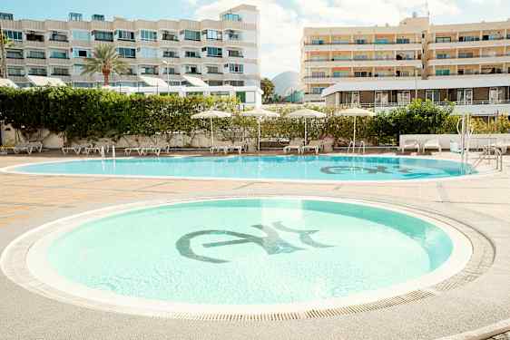 To swimmingpools omgivet af liggestole og parasoller, med hotelbygninger i baggrunden. (AI-genereret tekst)