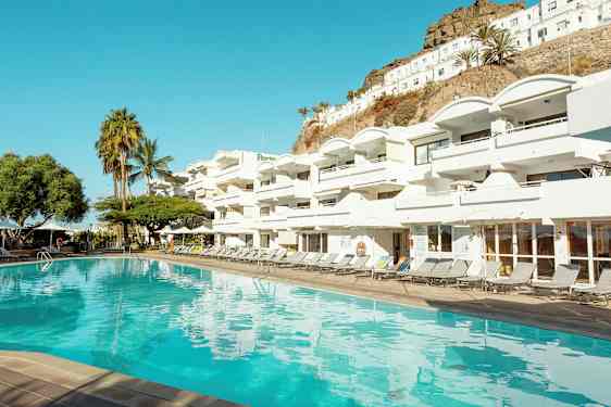 En pool foran et hvidt hotelkompleks med flere altaner. I baggrunden ses palmer og en skråning. (AI-genereret tekst)