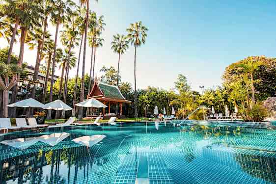 En swimmingpool omgivet af liggestole, parasoller og palmer med en thailandsk pavillon i baggrunden. (AI-genereret tekst)