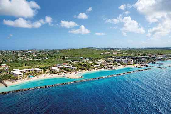 Luftfoto af en kystlinje med hotelbygninger ved en strand og grønne områder i baggrunden. (AI-genereret tekst)