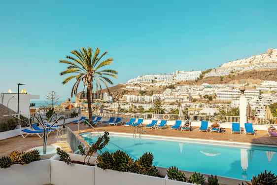 Udendørspool med liggestole og palmer samt udsigt over bjerge og bygninger. (AI-genereret tekst)