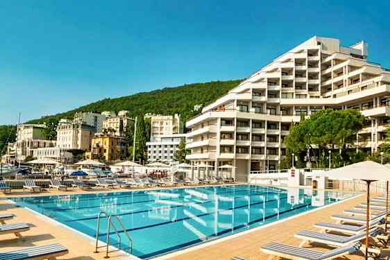 Poolområde med liggestole foran et hotel, omgivet af grønne bakker og flere bygninger i baggrunden. (AI-genereret tekst)