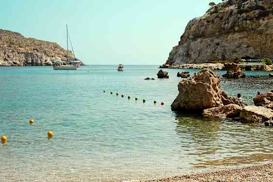 En bugt med klart vand og stenede kystlinje, hvor en båd og et mindre fartøj ses i baggrunden. (AI-genereret tekst)