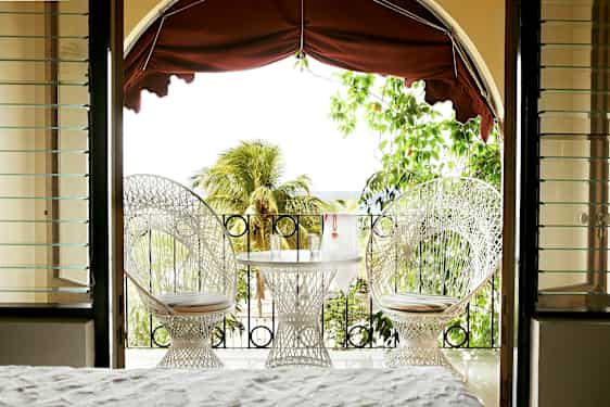 Udsigt fra et rum mod en terrasse med to hvide stole og et bord med glas, omgivet af grønt og palmer. (AI-genereret tekst)